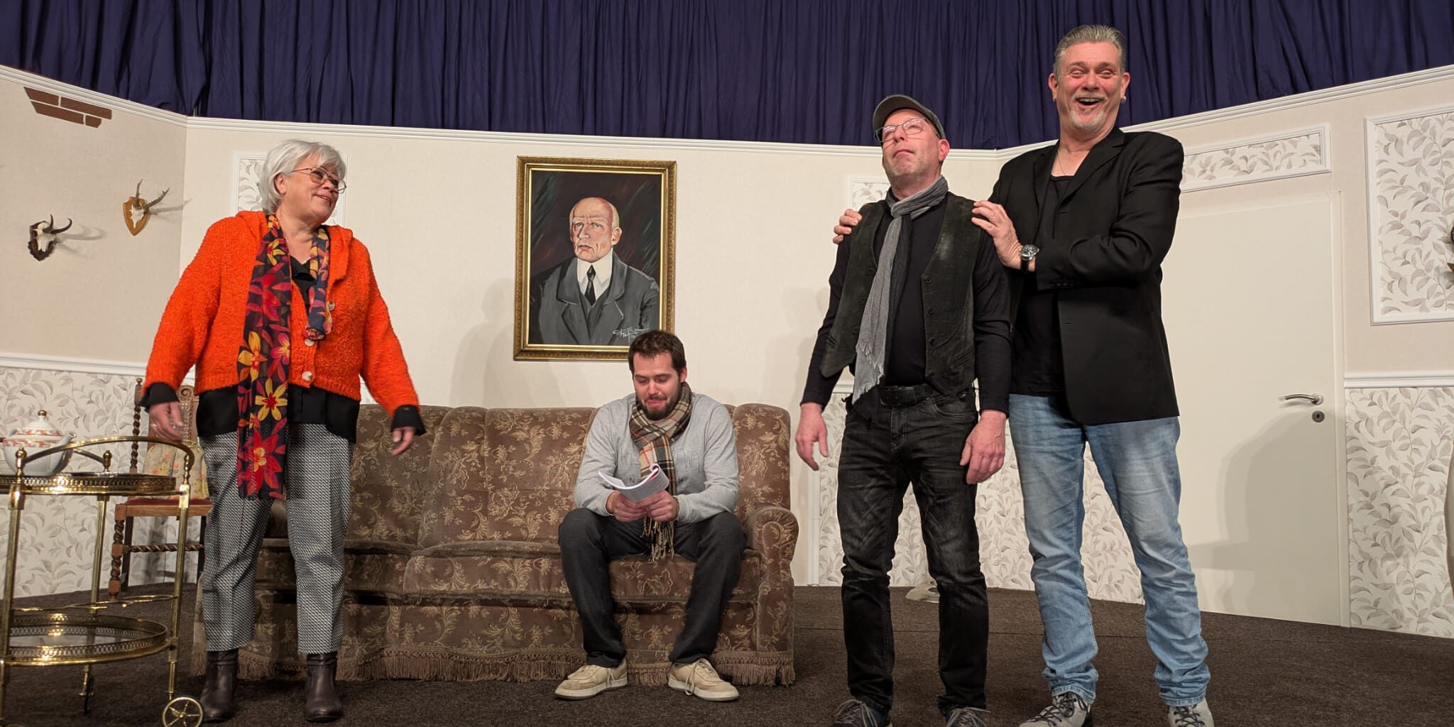 Vier Schauspieler spielen auf einer Theaterbühne in einem Wohnzimmer-Setting mit Sofa und Portrait.