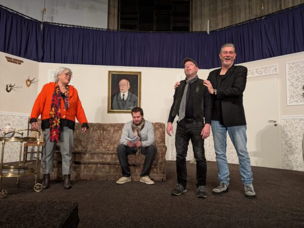 Foto: Freies Theater Auf Draht Vier Schauspieler spielen auf einer Theaterbühne in einem Wohnzimmer-Setting mit Sofa und Portrait.