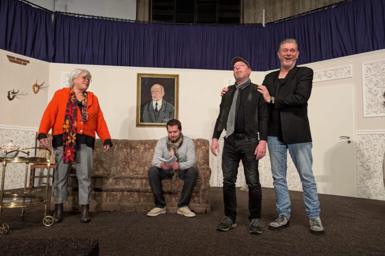 Vier Schauspieler spielen auf einer Theaterbühne in einem Wohnzimmer-Setting mit Sofa und Portrait.