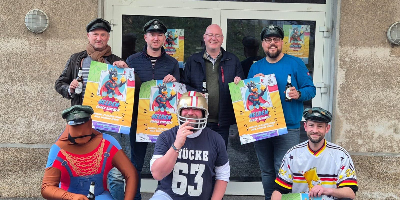 Sieben Männer vor einer Eingangstür, sie halten Werbeplakate in der Hand für die Motto-Party "Helden deiner Kindheit"
