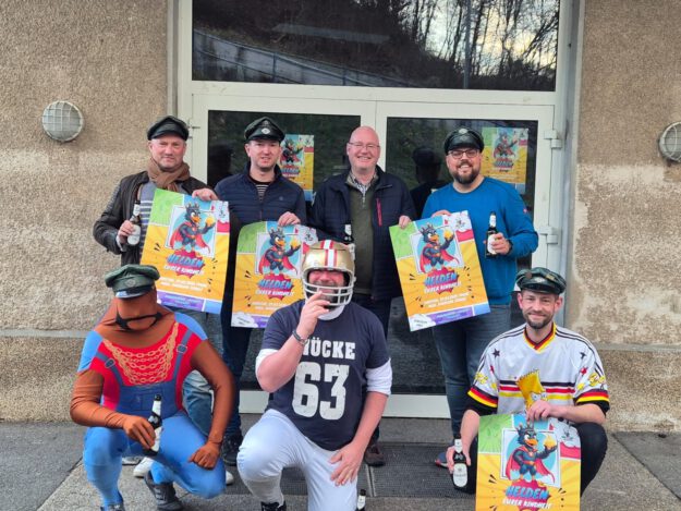 Foto: Kompanie Kelleramt Sieben Männer vor einer Eingangstür, sie halten Werbeplakate in der Hand für die Motto-Party "Helden deiner Kindheit"