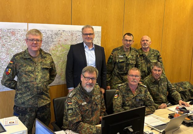 Sechs Bundeswehrsoldaten und eine Person im Anzug stehen vor mehreren PC-Bildschrimen. Im Hintergrund eine Landkarte.