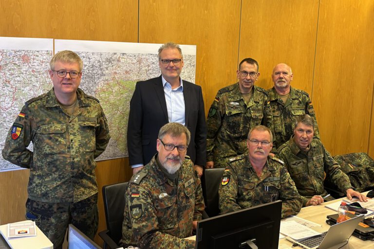 Sechs Bundeswehrsoldaten und eine Person im Anzug stehen vor mehreren PC-Bildschrimen. Im Hintergrund eine Landkarte.