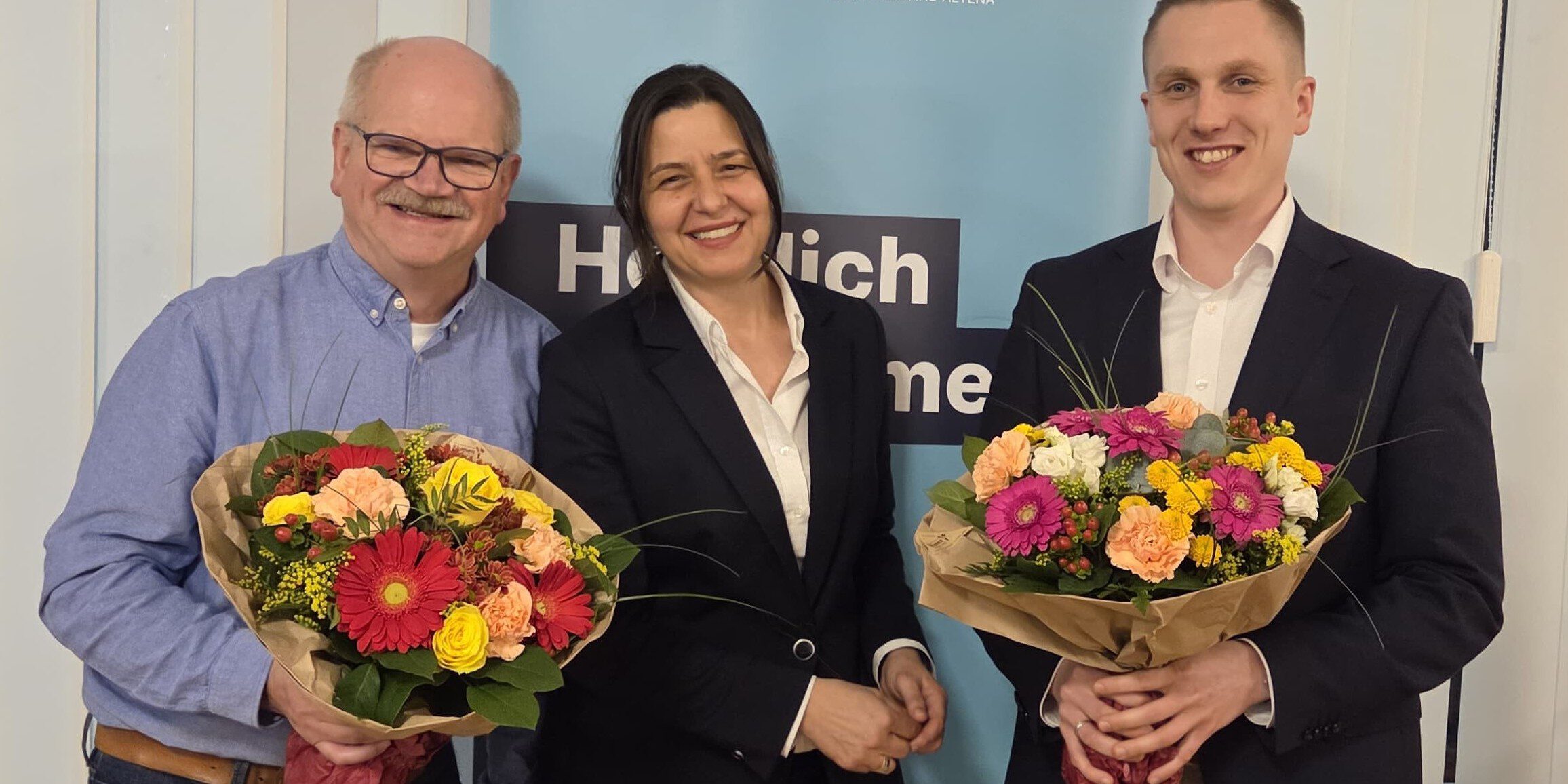 Politiker stehen vor einem Werbeplakat, zwei Männer halten Blumensträuße in der Hand.