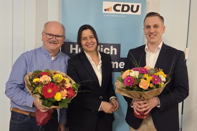Politiker stehen vor einem Werbeplakat, zwei Männer halten Blumensträuße in der Hand.
