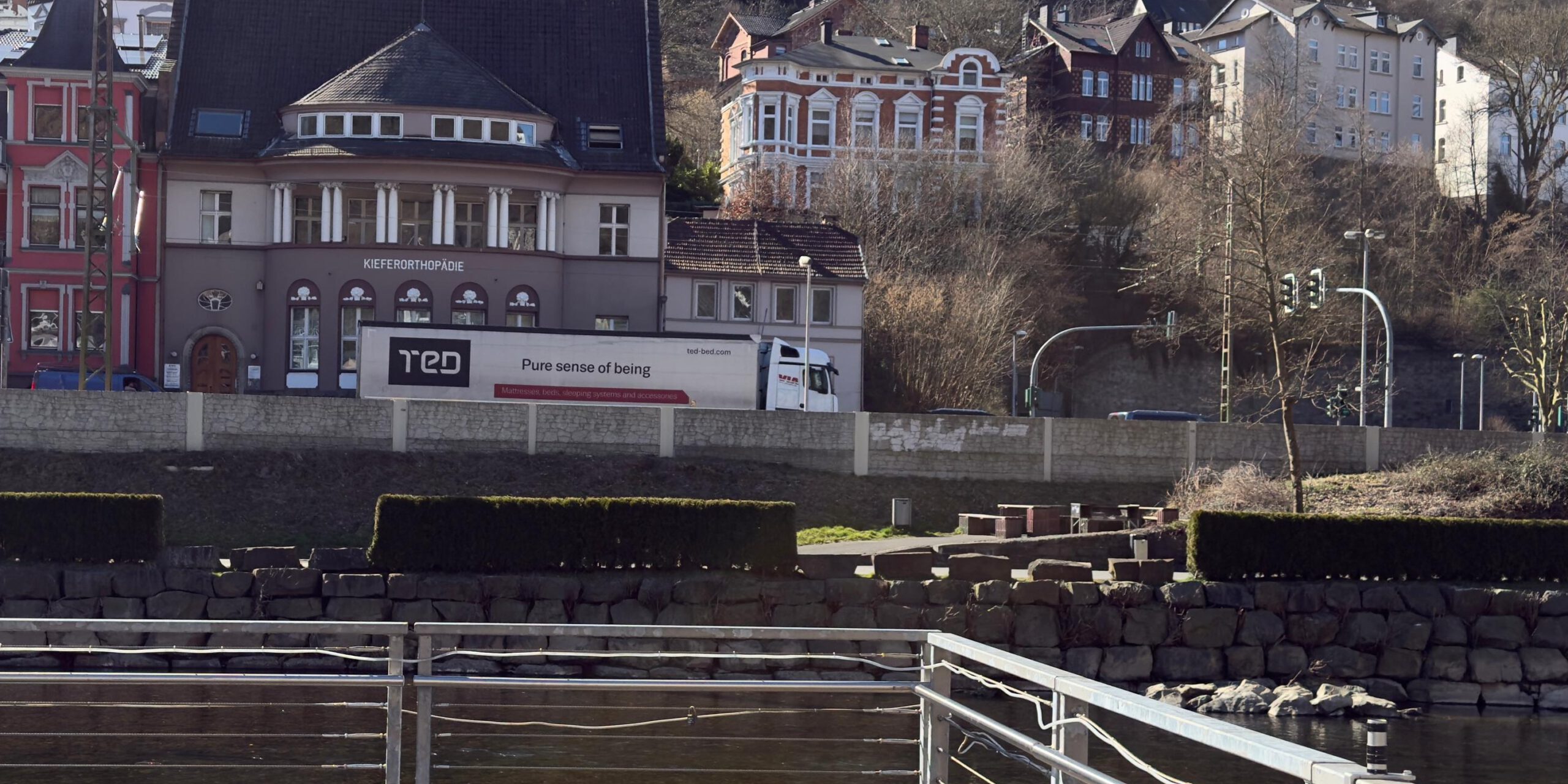 Ein Lkw fährt auf einer Straße vor mehreren Häusern. Im Vordergrund eine Hecke.