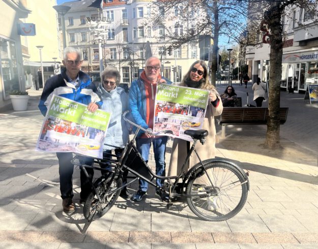 Vier Personen stehen in der Innenstadt und halten Werbeplakate für den Hollandmarkt in den Händen.
