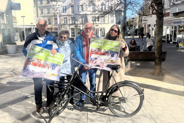 Vier Personen stehen in der Innenstadt und halten Werbeplakate für den Hollandmarkt in den Händen.