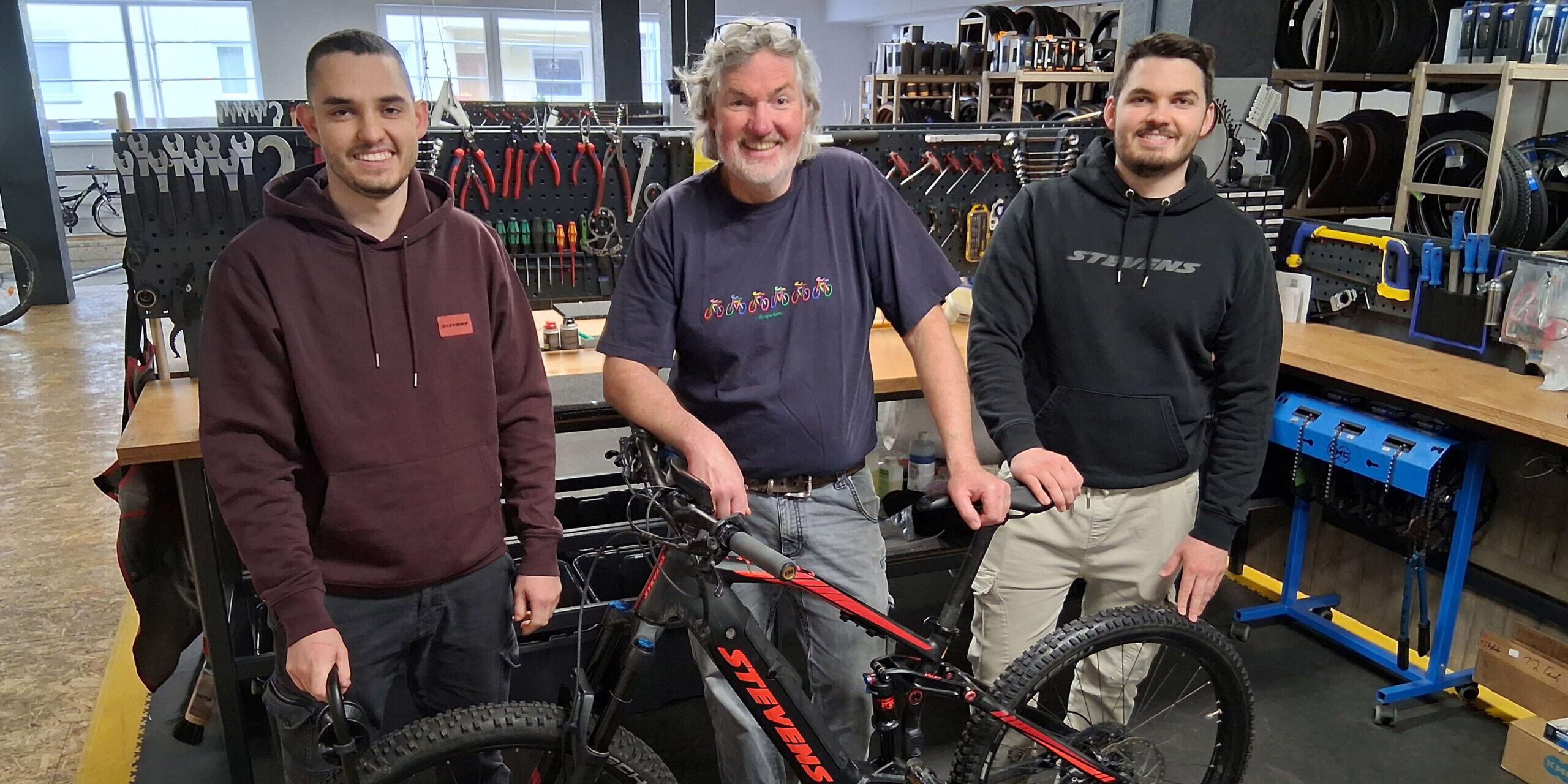 Drei Männer stehen in einer Fahrradwerkstatt vor einem Fahrrad.
