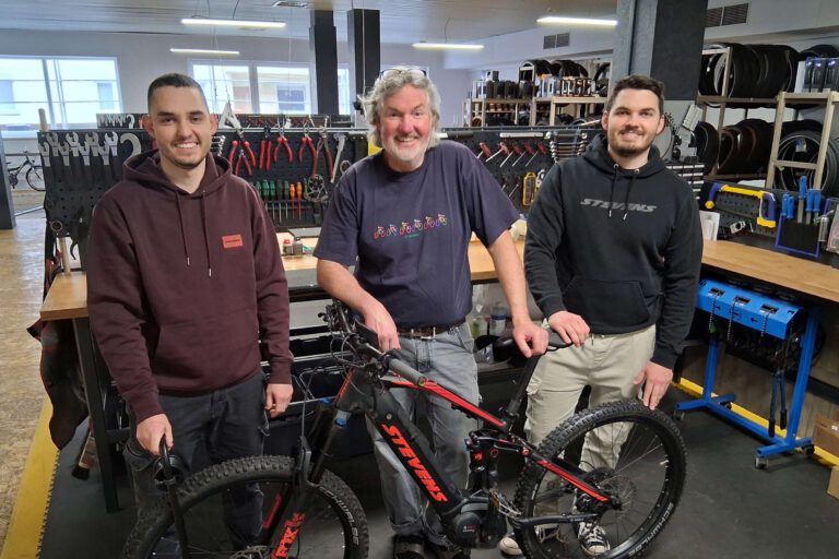 Drei Männer stehen in einer Fahrradwerkstatt vor einem Fahrrad.