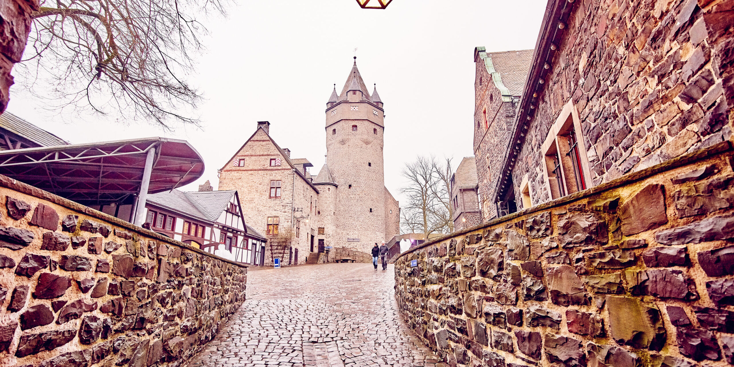 Die Jugendherberge auf Burg Altena war die erste ständige Jugendherberge der Welt.
© Foto: DJH Pflasterweg führt durch ein Burggelände mit historischen Stein- und Fachwerkgebäuden zur Burg Altena, im Hintergrund der markante Burgturm, vorne eine leuchtende Laterne.“