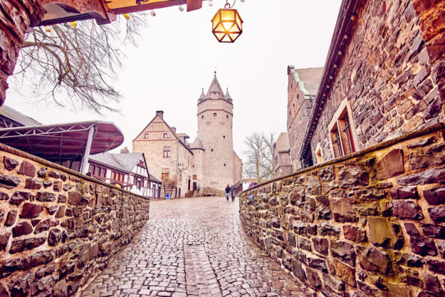Foto: DJH Pflasterweg führt durch ein Burggelände mit historischen Stein- und Fachwerkgebäuden zur Burg Altena, im Hintergrund der markante Burgturm, vorne eine leuchtende Laterne.“