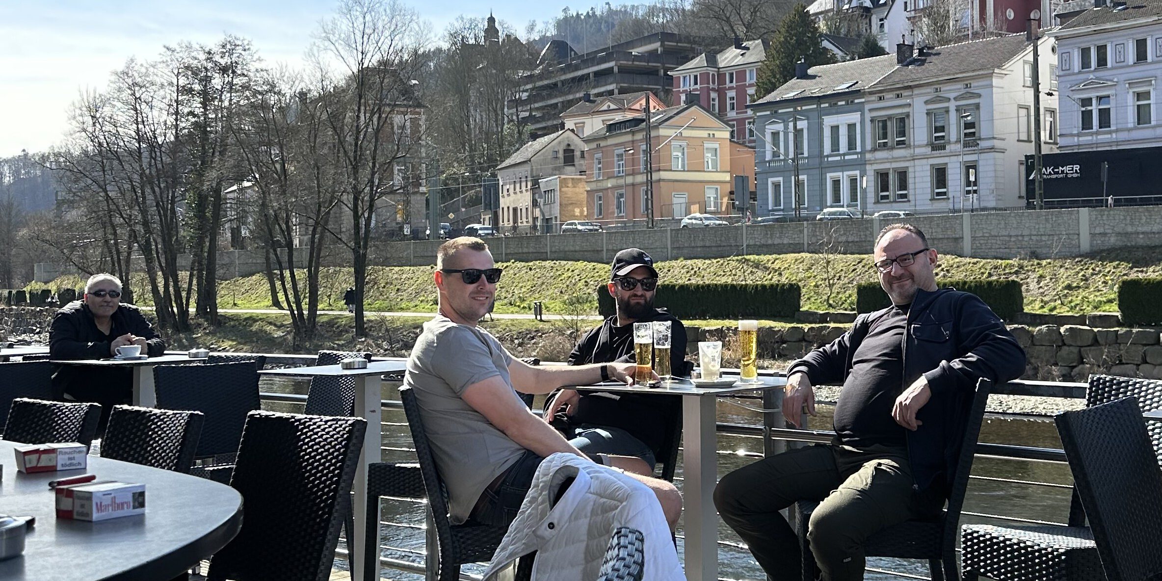 Vier Männer sitzen auf einer Terrasse, trinken Bier. Im Hintergrund fließt ein Fluss.