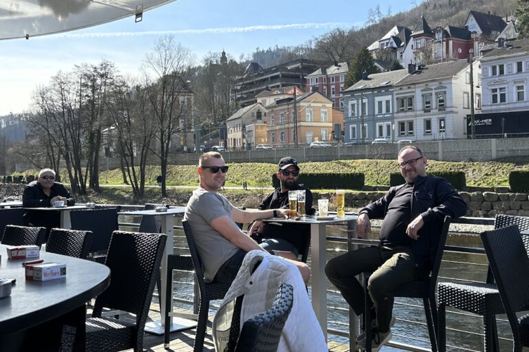 Vier Männer sitzen auf einer Terrasse, trinken Bier. Im Hintergrund fließt ein Fluss.