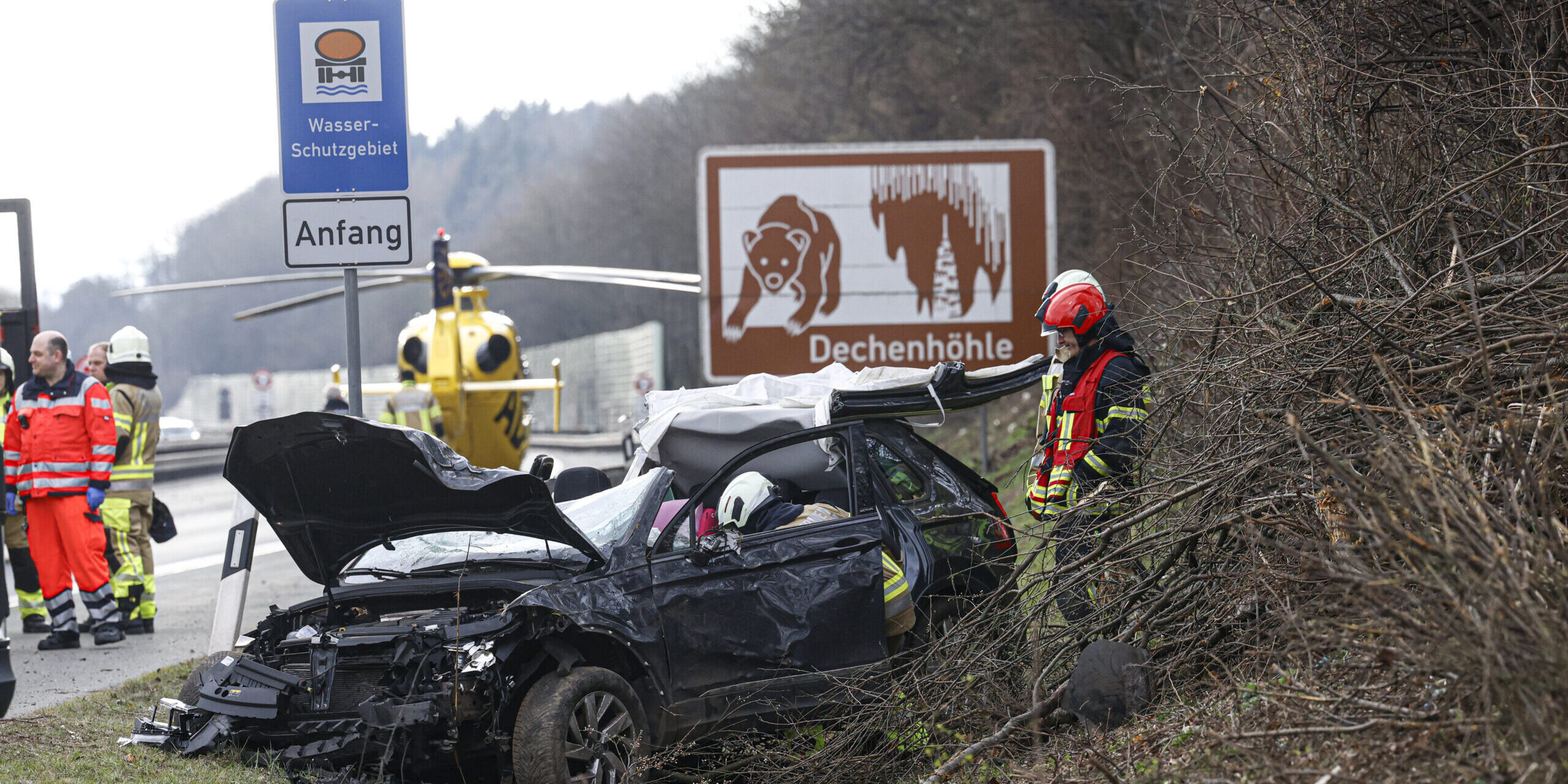 Ein zerstörter schwarzer Pkw auf der Autobahn. Einsatzkräfte stehen drumherum, im Hintergrund ist ein Rettungshubschrauber gelandet.