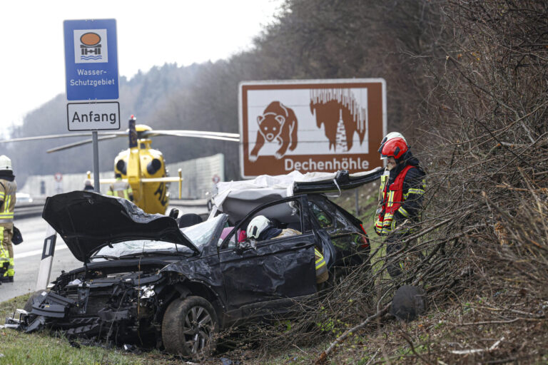 Ein zerstörter schwarzer Pkw auf der Autobahn. Einsatzkräfte stehen drumherum, im Hintergrund ist ein Rettungshubschrauber gelandet.