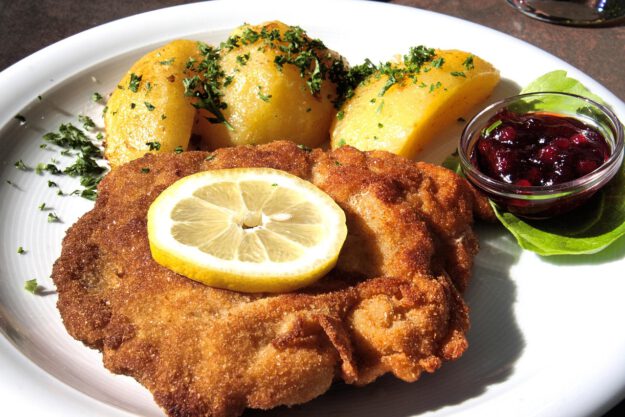 Foto: Reinhard Thrainer/pixabay Panierte Schnitzel auf einem weißen Teller, serviert mit Zitronenscheibe und Petersilie, daneben ein kleiner Salat und Kartoffeln.