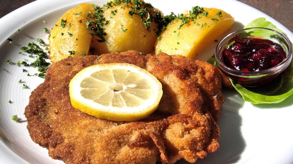 Panierte Schnitzel auf einem weißen Teller, serviert mit Zitronenscheibe und Petersilie, daneben ein kleiner Salat und Kartoffeln.