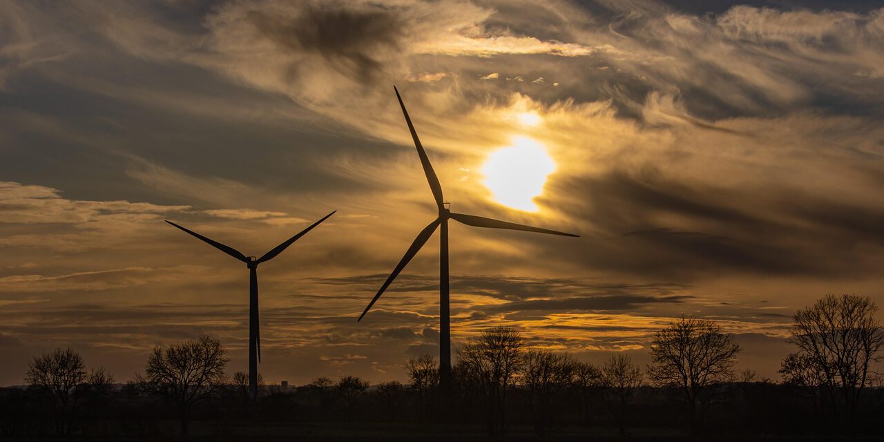 Zwei Windräder im Sonnenuntergang vor einem Wald.