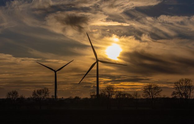 Foto: TheOtherKev/pixabay Zwei Windräder im Sonnenuntergang vor einem Wald.