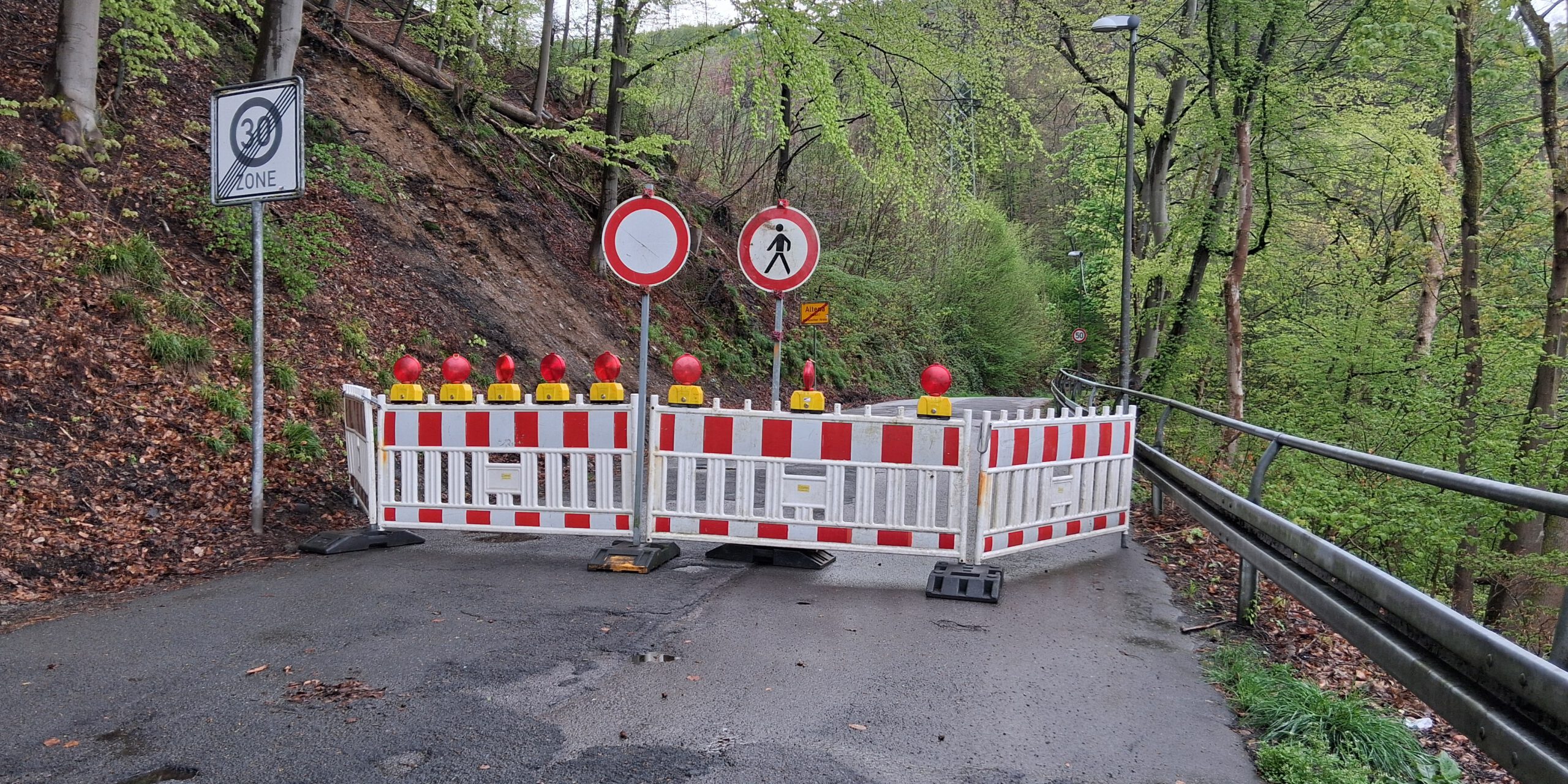 Die Straße Am Knerling musste zwischen Ortsausgang Altena und Opperhusen kurzfristig voll gesperrt werden. Ein Felsbrocken war auf die Fahrbahn gestürzt.
© Foto: Ilka Kremer Absperrbaken blockieren eine Straße. Schilder verbieten die Durchfahrt für Autos und Fußgänger.