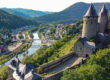 Ansicht der Burg Altena auf einem bewaldeten Hügel im Sommer. Unterhalb fließt der Fluss Lenne.