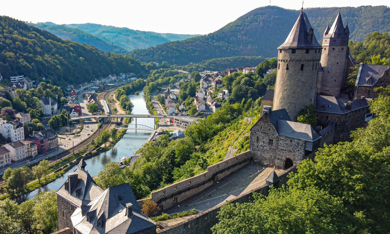 Ansicht der Burg Altena auf einem bewaldeten Hügel im Sommer. Unterhalb fließt der Fluss Lenne.