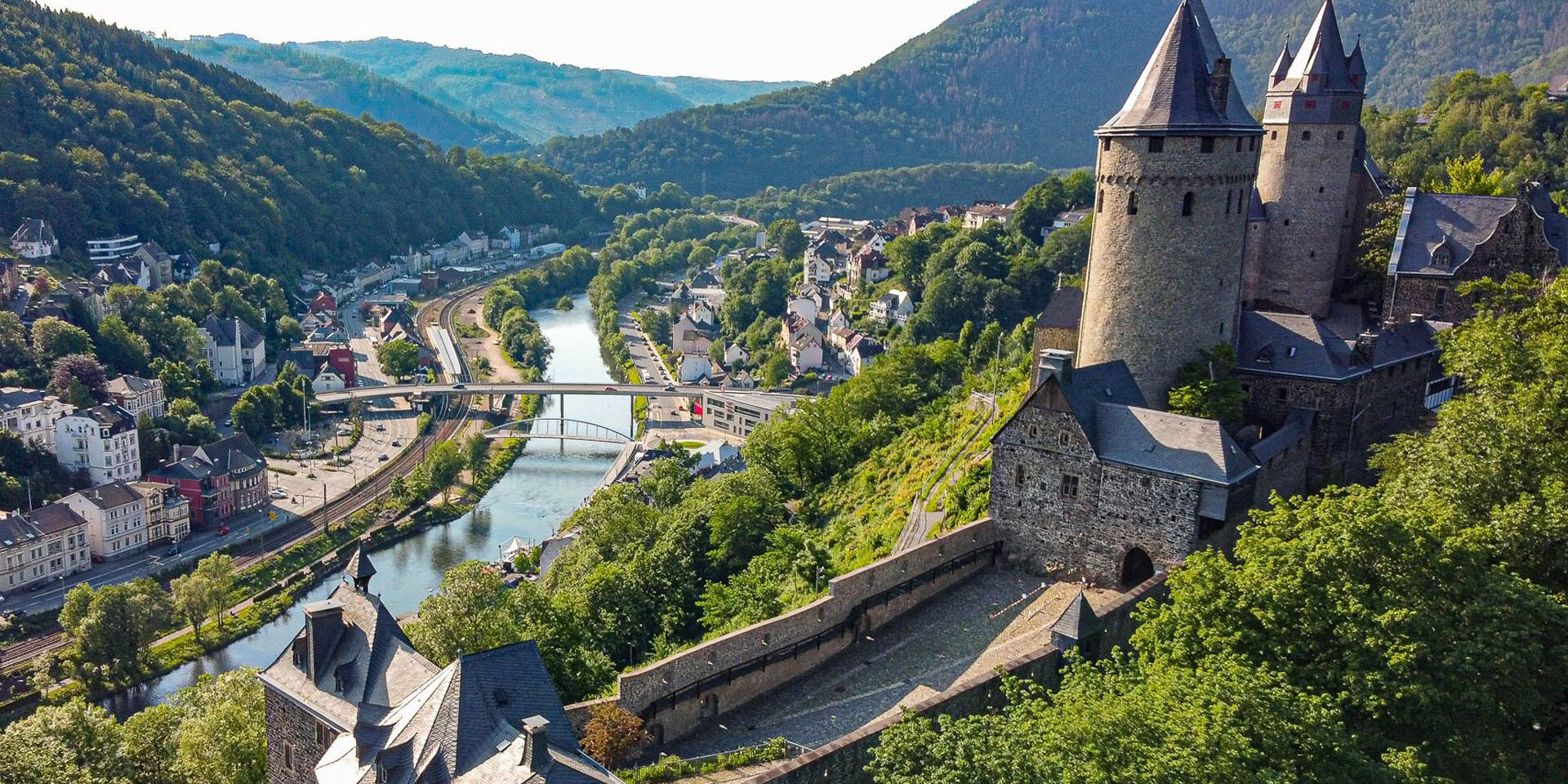 Ansicht der Burg Altena auf einem bewaldeten Hügel im Sommer. Unterhalb fließt der Fluss Lenne.