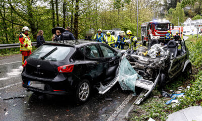 Zwei zerstörte Autos stehen nach einem Unfall auf der Straße. Im Hintergrund steht ein Feuerwehrfahrzeug und Rettungswagen. Einsatzkräfte von Polizei und Feuerwehr sind sichtbar.
