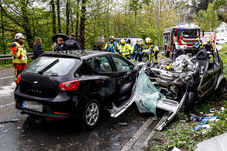 Zwei zerstörte Autos stehen nach einem Unfall auf der Straße. Im Hintergrund steht ein Feuerwehrfahrzeug und Rettungswagen. Einsatzkräfte von Polizei und Feuerwehr sind sichtbar.