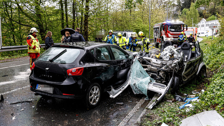 Zwei zerstörte Autos stehen nach einem Unfall auf der Straße. Im Hintergrund steht ein Feuerwehrfahrzeug und Rettungswagen. Einsatzkräfte von Polizei und Feuerwehr sind sichtbar.