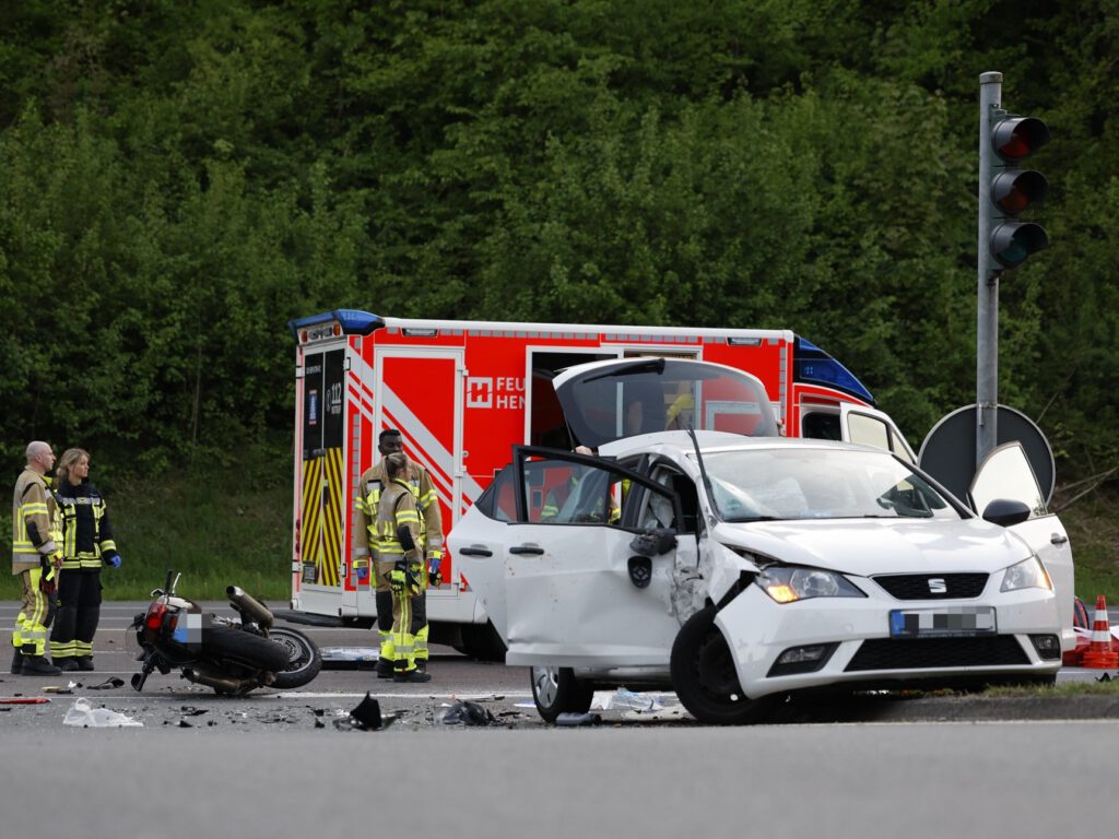 Ein an der Fahrerseite erheblich beschädigtes weißes Auto steht an einer Verkehrsinsel mit offenen Türen. Im Hintergrund liegt ein Motorrad auf der Straße, daneben stehen drei Rettungskräfte. Ganz hinten steht ein Rettungswagen.