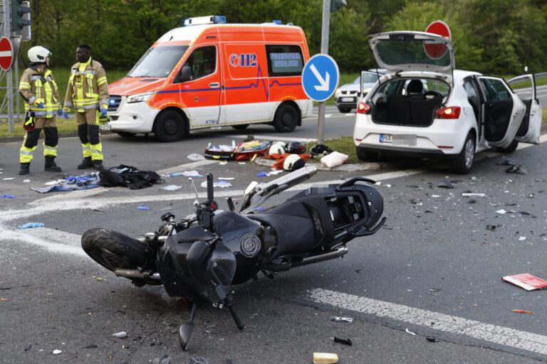 Das Wrack eines Motorrads liegt nach einem Unfall auf der Straße. Im Hintergrund stehen eine weißer Pkw und ein Notarztfahrzeug. Links am Bildrand drei Rettungskräfte.