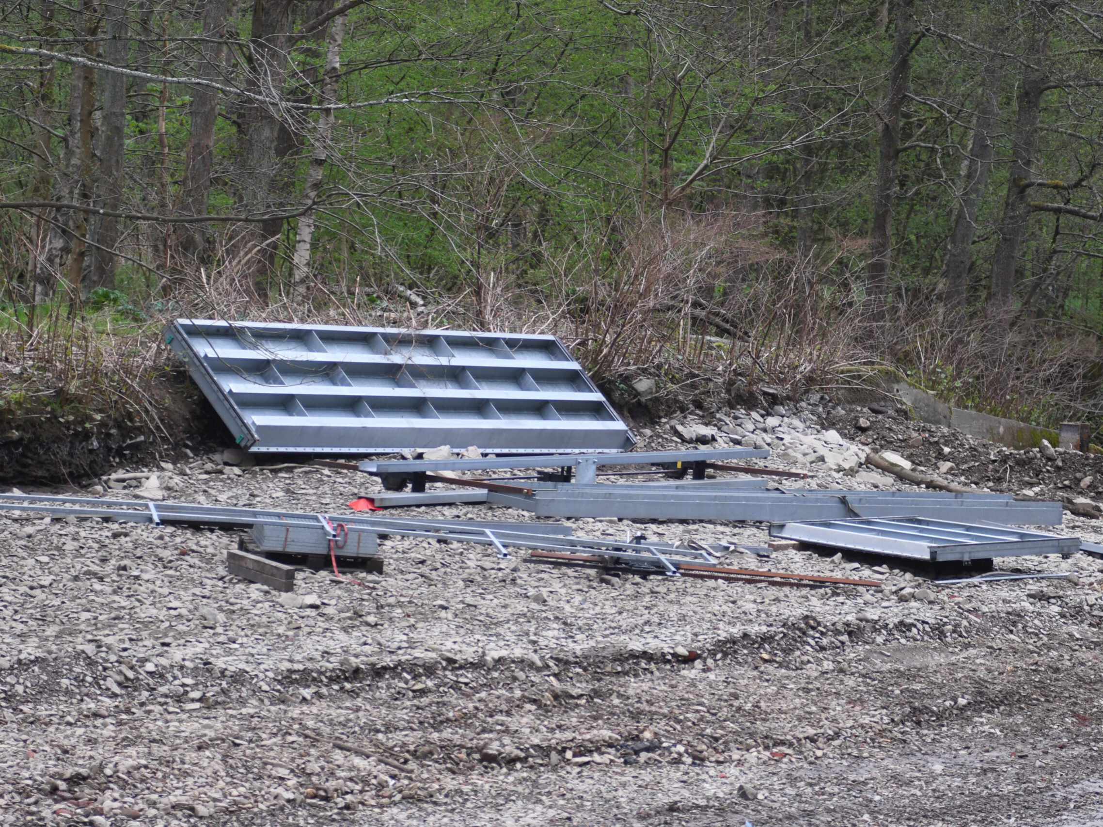 Metallteile liegen am Flussufer vor einer bewaldeten Böschung.