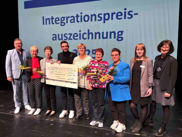 Foto: Märkischer Kreis Eine Gruppe Menschen steht nach einer Preisverleihung auf einer Bühne und hält einen 500 Euro Spendenscheck in der Hand.