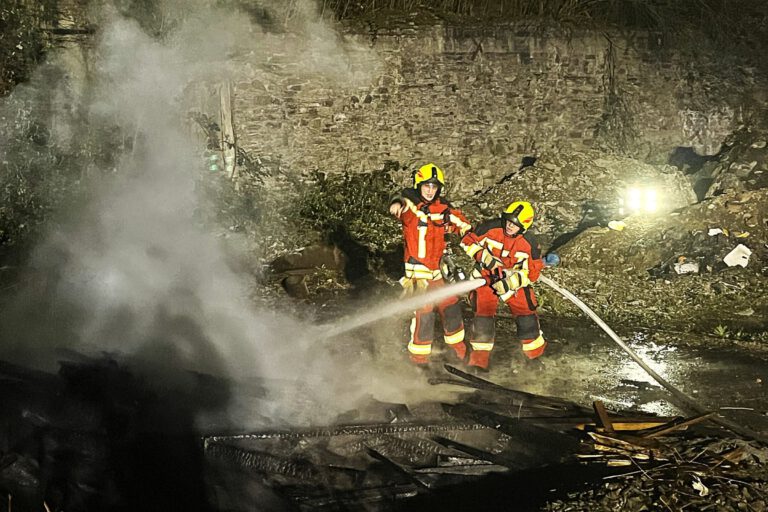 Zwei Einsatzkräfte der Feuerwehr stehen bei Dunkelheit vor einem brennenden Holzhaufen.