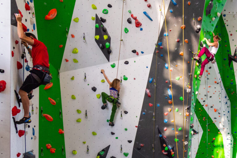 Drei Personen beim Klettern an einer bunten Indoor-Kletterwand mit farbigen Griffen und Sicherungsseilen; ein Kind klettert in der Mitte zwischen zwei Erwachsenen.
