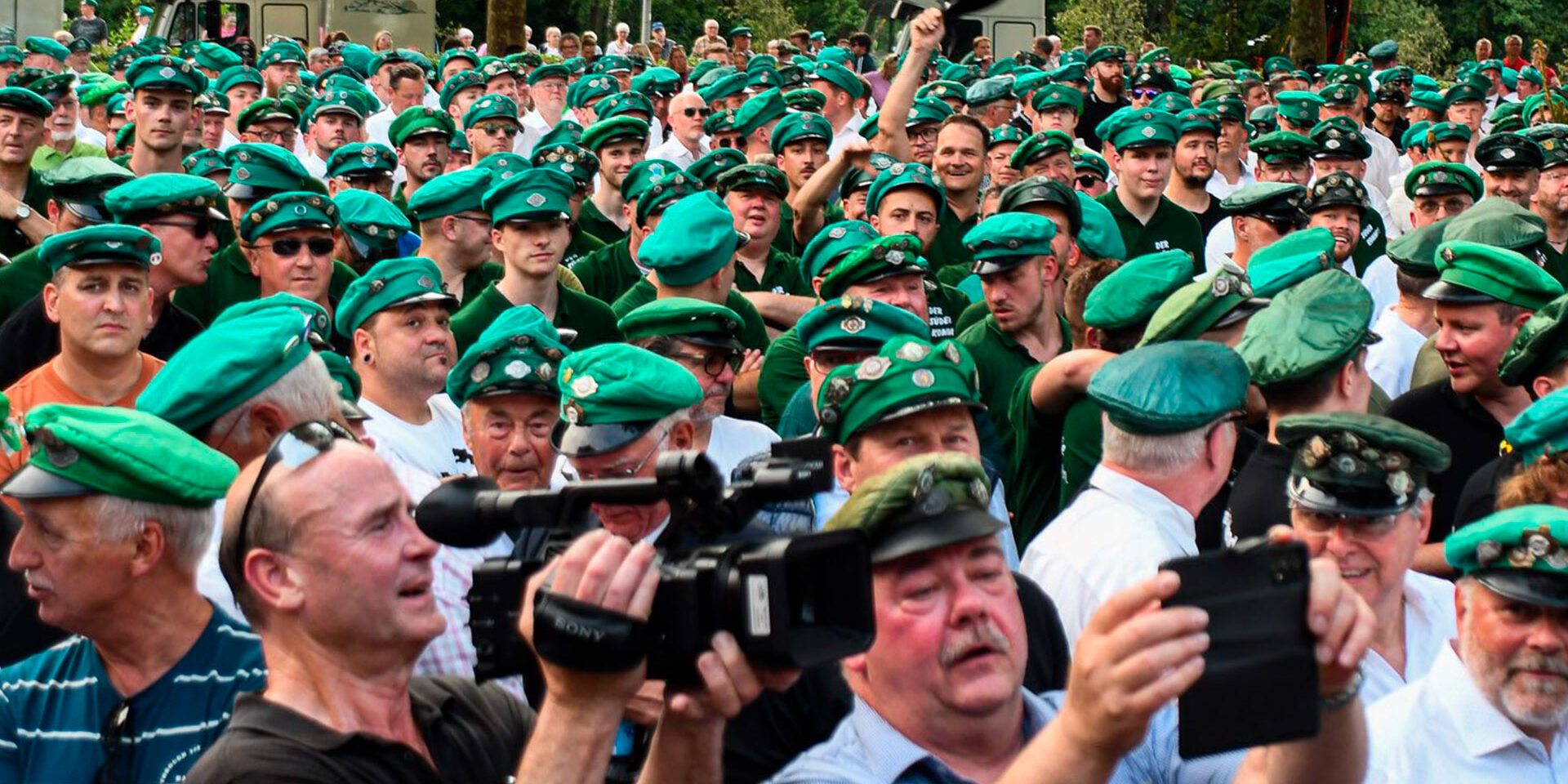 Menschenmenge bei einem Schützenfest, viele Besucher sitzen an langen Tischen im Zelt, einige tragen traditionelle Schützenuniformen mit grünen Mützen.
