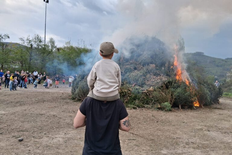 Ein Mann mit einem Kind auf dem Arm steht vor einem Osterfeuer und beobachtet dieses. Im Hintergrund stehen weitere Besucher um das Feuer herum.