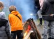 Mehrere Kinder stehen bei einem Osterfeuer im Freien. Sie tragen warme Kleidung und rösten Marshmallows über dem offenen Feuer aus großen Holzscheiten. Im Hintergrund sind weitere Menschen zu sehen, dichter Rauch steigt auf.
