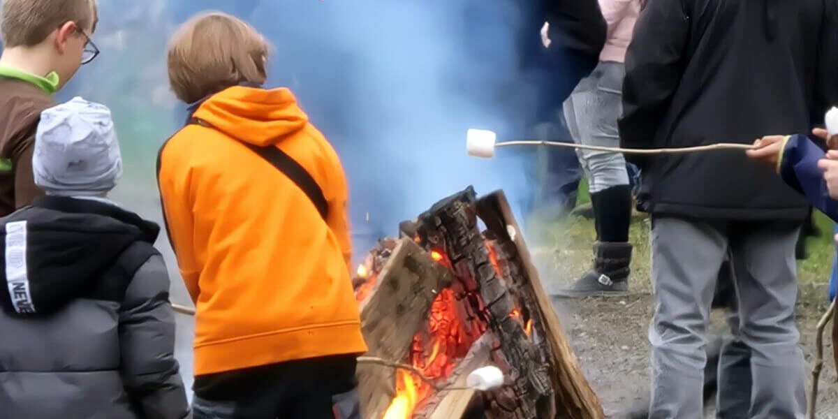 Mehrere Kinder stehen bei einem Osterfeuer im Freien. Sie tragen warme Kleidung und rösten Marshmallows über dem offenen Feuer aus großen Holzscheiten. Im Hintergrund sind weitere Menschen zu sehen, dichter Rauch steigt auf.