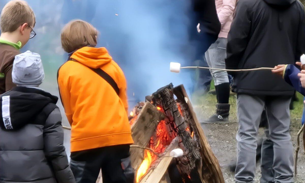 Mehrere Kinder stehen bei einem Osterfeuer im Freien. Sie tragen warme Kleidung und rösten Marshmallows über dem offenen Feuer aus großen Holzscheiten. Im Hintergrund sind weitere Menschen zu sehen, dichter Rauch steigt auf.