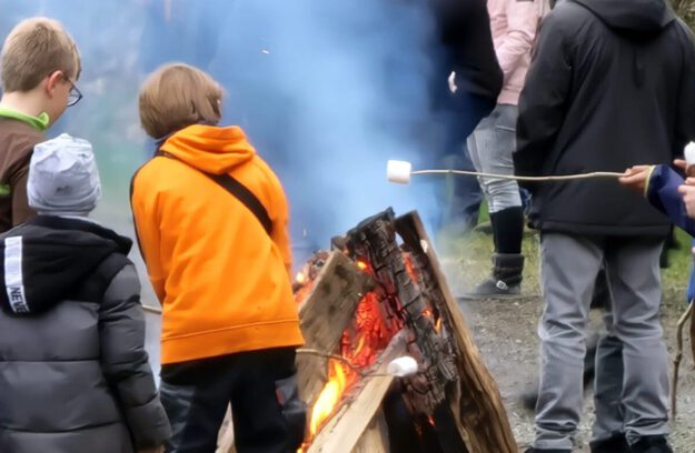 Mehrere Kinder stehen bei einem Osterfeuer im Freien. Sie tragen warme Kleidung und rösten Marshmallows über dem offenen Feuer aus großen Holzscheiten. Im Hintergrund sind weitere Menschen zu sehen, dichter Rauch steigt auf.