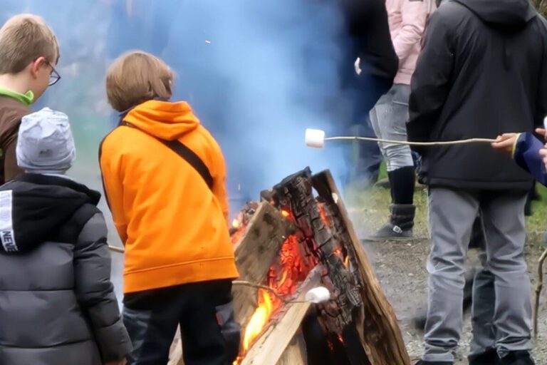 Mehrere Kinder stehen bei einem Osterfeuer im Freien. Sie tragen warme Kleidung und rösten Marshmallows über dem offenen Feuer aus großen Holzscheiten. Im Hintergrund sind weitere Menschen zu sehen, dichter Rauch steigt auf.