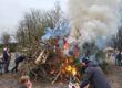 Osterfeuer mit brennendem Holzhaufen aus Ästen und Baumstämmen, dichter Rauch steigt auf, mehrere Menschen, darunter Kinder, beobachten das Feuer.