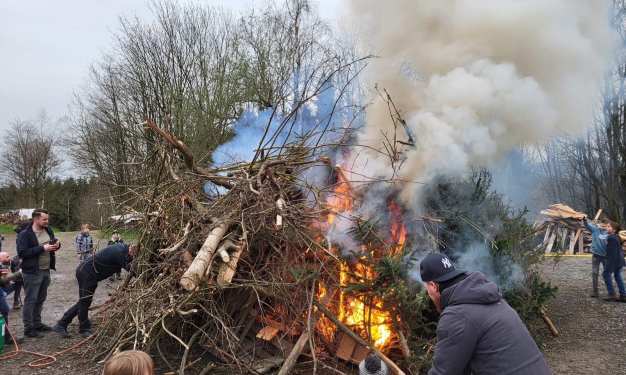 Osterfeuer mit brennendem Holzhaufen aus Ästen und Baumstämmen, dichter Rauch steigt auf, mehrere Menschen, darunter Kinder, beobachten das Feuer.
