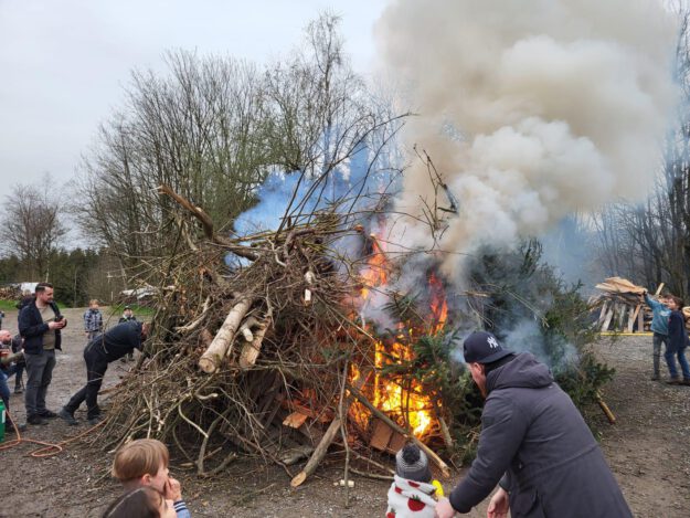 Archivfoto: Ilka Kremer Osterfeuer mit brennendem Holzhaufen aus Ästen und Baumstämmen, dichter Rauch steigt auf, mehrere Menschen, darunter Kinder, beobachten das Feuer.