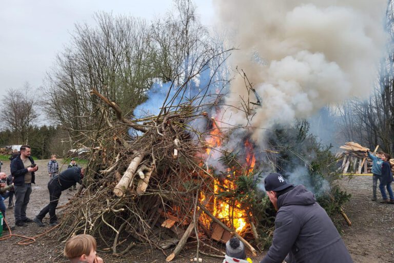 Archivfoto: Ilka Kremer Osterfeuer mit brennendem Holzhaufen aus Ästen und Baumstämmen, dichter Rauch steigt auf, mehrere Menschen, darunter Kinder, beobachten das Feuer.