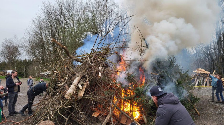 Osterfeuer mit brennendem Holzhaufen aus Ästen und Baumstämmen, dichter Rauch steigt auf, mehrere Menschen, darunter Kinder, beobachten das Feuer.
