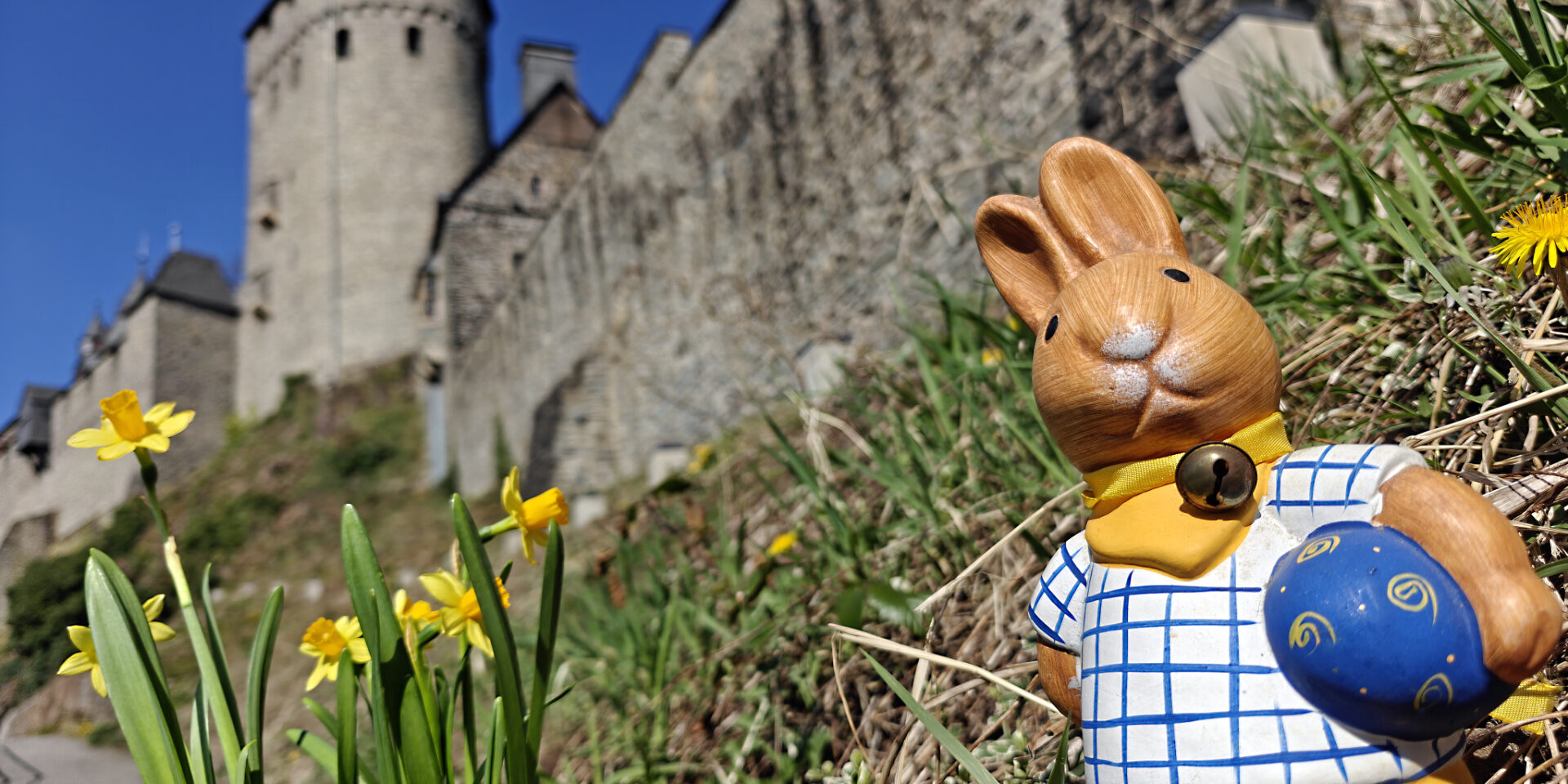 Eine Osterhasen Figur steht vor den Mauern der Burg Altena, daneben blühen Blumen.